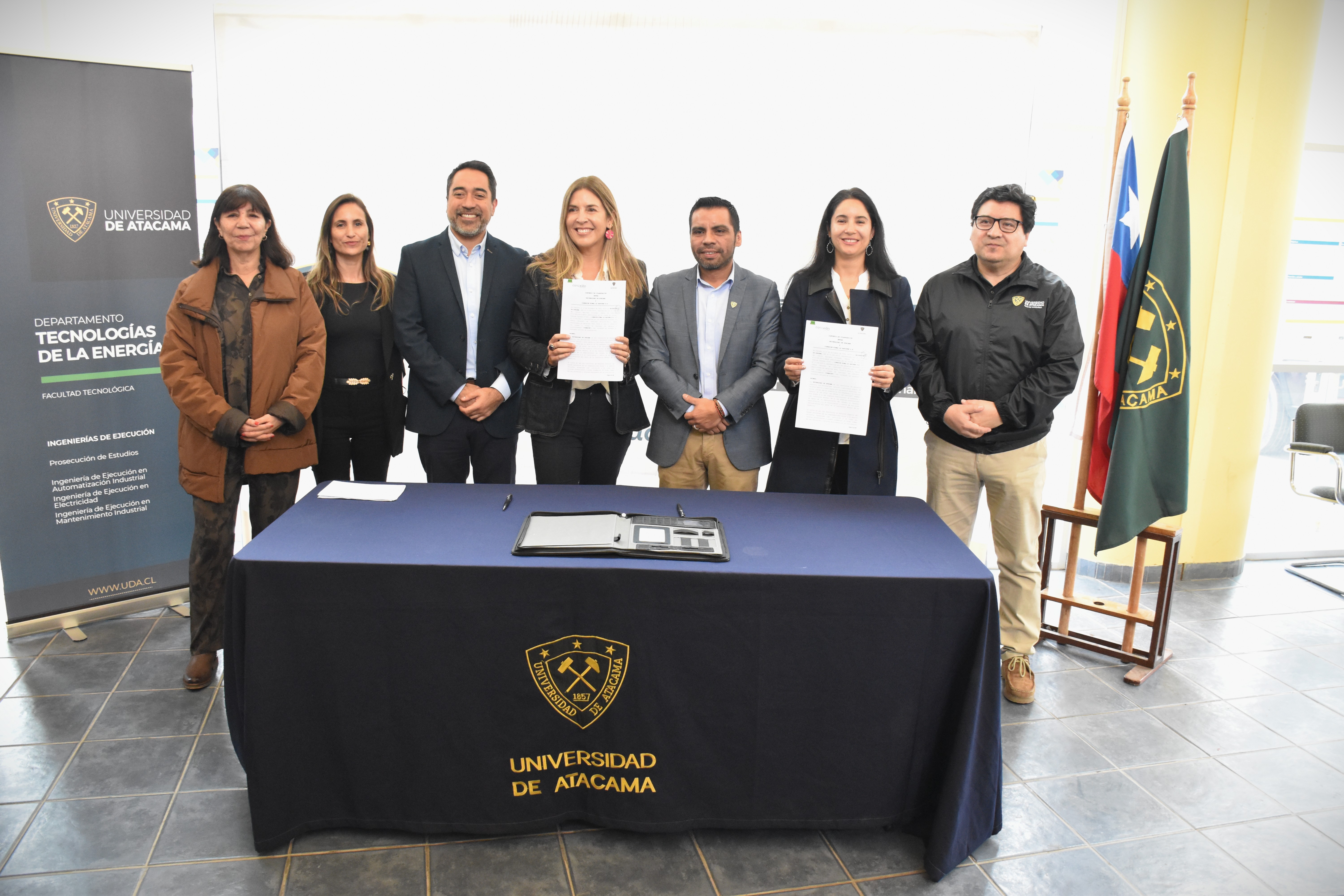 Siete personas de pie en el centro con una mesa en frente, un pendón a la izquierda y dos banderas institucionales a la derecha