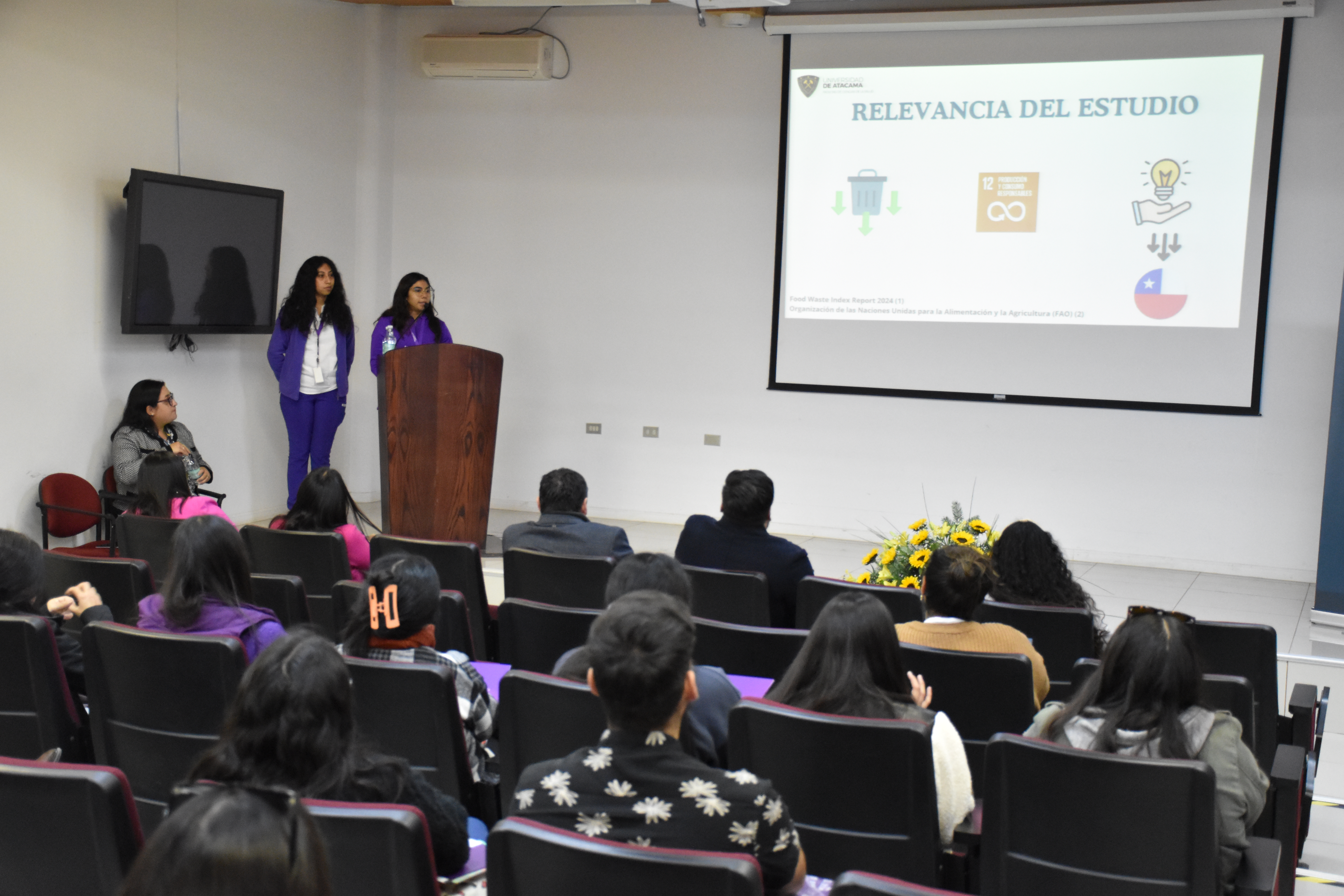 Dos mujeres a la izquierda en una tarima frente a un podio, exponiendo junto a una gran proyección en fondo blanco. En la mitad inferior, un público sentado mirando a la exposición