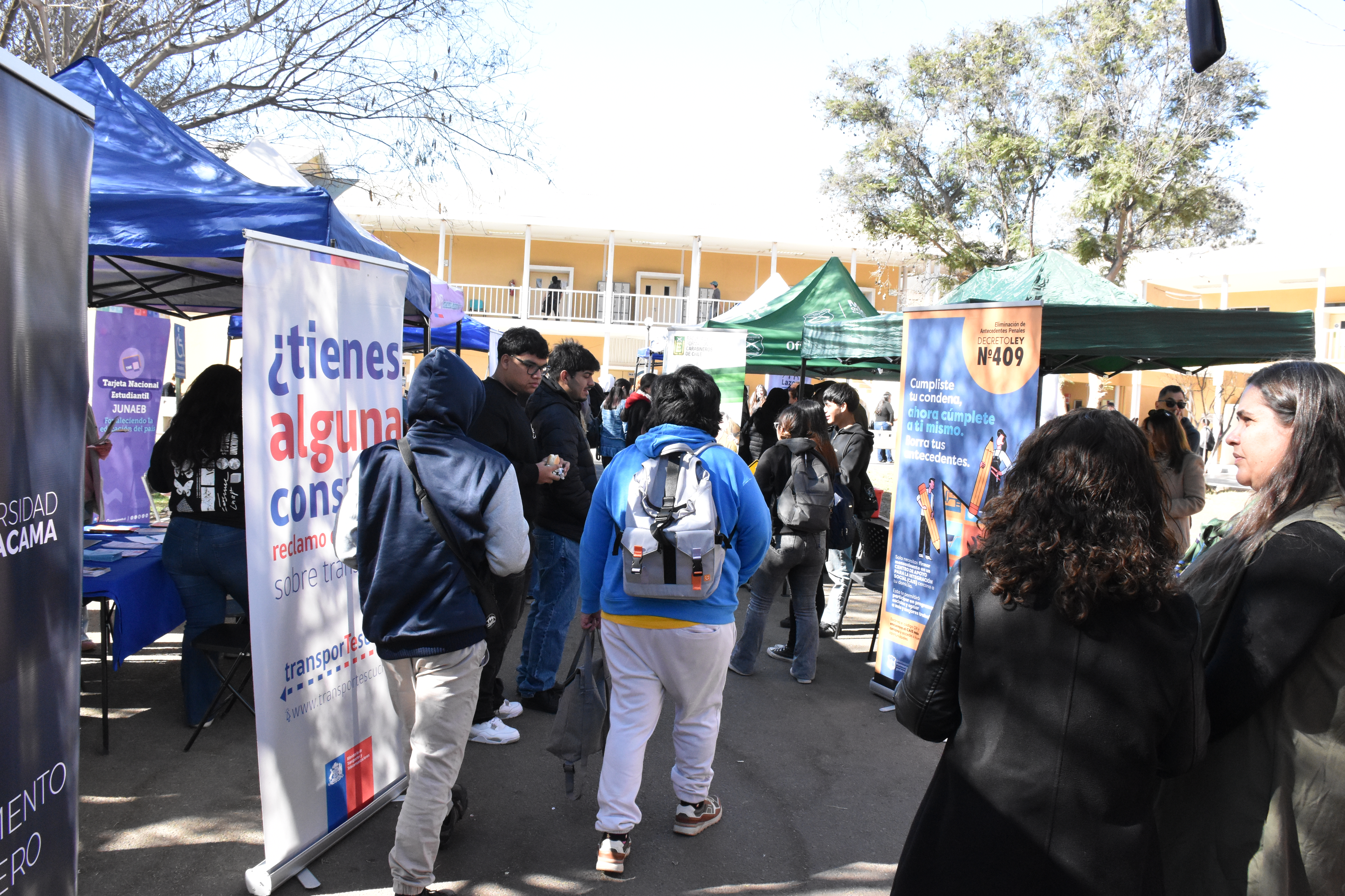 Feria de Seguridad UDA en el Campus Sur. Gente caminando en linea en el centro, a los costados hay toldos y pendones de instituciones de seguridad del gobierno de Chile