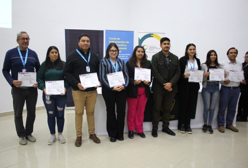 Personas certificadas por el curso de gobernanza junto a sus profesores, de pie y sosteniendo sus diplomas frente a tres pendones institucionales. Pared blanca y suelo gris